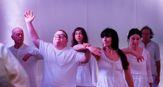 A group of 6 people standing in a line, wearing white. They are leaning on each others shoulders and the second person from the left is holding their arm up and looking towards the ceiling. 