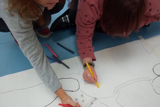 Two people are seated on the floor drawing on a large piece of paper. The floor is blue. One is drawing a necklace on the shape of a hand and the other is drawing a decoration on the shape of a jumper.