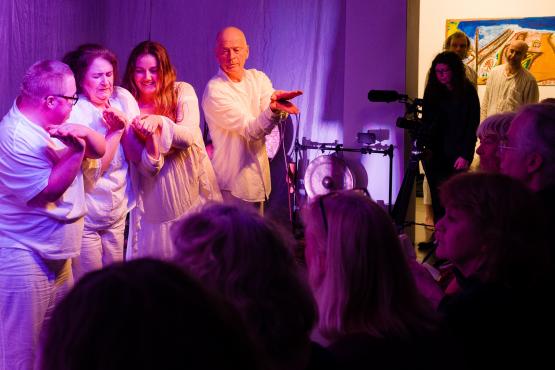 Four dancers in white dance in front of a white background facing a seated audience. The image shows the backs of the audience members' heads. The room is smallish and dark except for the purplish white light on the dancers and we can see an opening with a painting on the wall outside. In the opening a person is filming the performance.