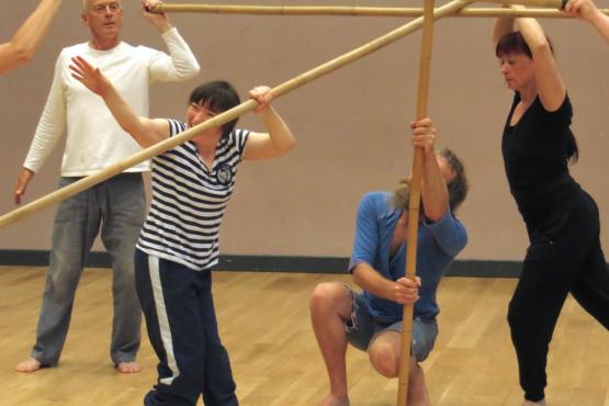 Four people are dancing in a rehearsal studio. Three of them are holding up thick bamboo poles. The poles support the fourth dancer to stand.