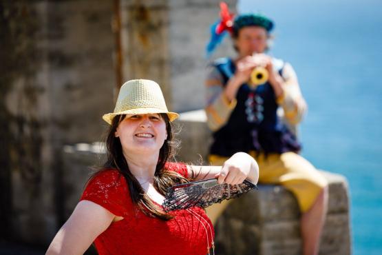 2 people performing on stage. The person standing in the foreground is wearing a red top and straw hat and smiling at the camera. The second person is sitting on a stone wall with the sea behind them playing a horn instrument. 