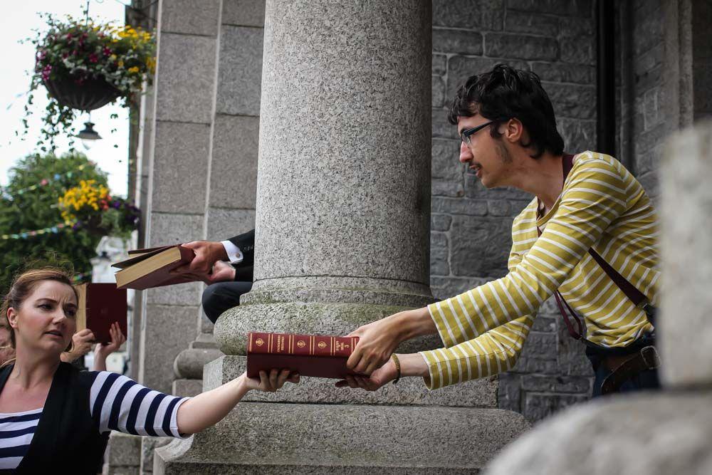 One dancer passes another dancer a book as part of the performance. They are outside dancing between big pillars.