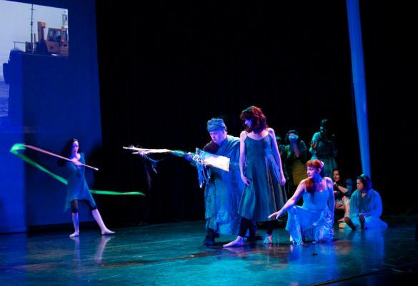 Eight dancers perform on stage with a projected image behind them. Three dancers are crouching or sitting and five are standing. One dancer is waving a  green streamer in an arc around their body. All the other dancers are looking to the left.