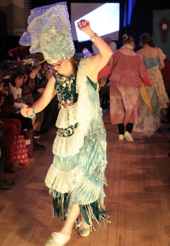 A person is dressed in a fashion show costume with a dark background. She is wearing a tall hat made of bubble wrap and a blue dress with a white fringe going round it in a spiral from top to bottom like waves on the sea.