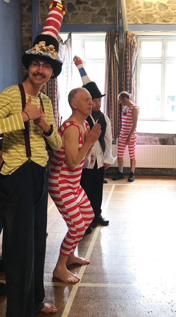 Four people are standing wearing fun dressing up costumes and hats in a light-filled rehearsal hall. The one nearest to the camera is wearing a yellow top and black trousers with braces and a hat with a lighthouse on top and he is putting both thumbs up and smiling.