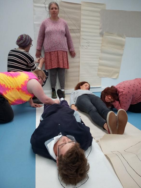 6 people taking part in a drawing workshop where they are drawing around each others body outline on large pieces of paper. 2 people are lying on the ground on top of paper. 2 people are knelt on the floor drawing around the outline of the others lying on the floor. 1 person is standing against paper hung on the wall while another person draws around their body shape.