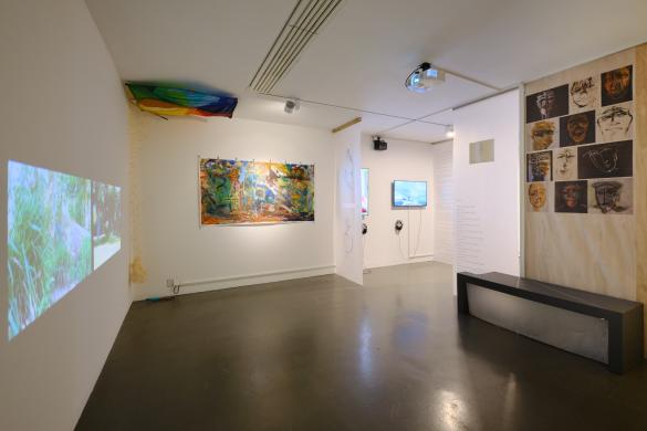 film projected on wall, kite on ceiling in corner, large  multi coloured painting, some monitors and words and brown portraits hung above a black benc