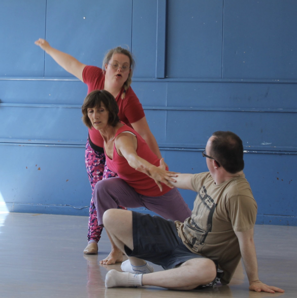 3 people dancing. They are holding hands in a line. 1 person is crouched on the floor holding hands with the middle person who is bent down. The third person is standing at the back holding the middle person by the hand and reaching out their other arm to the left