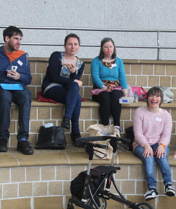 Four people sit eating their picnic lunches on some wide steps. They are all smiling. From left to right the person far left is eating a yoghurt, the next person is holding up a lunchbox, the two people on the right have their hands in their laps.