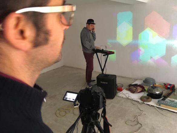 Two people are standing in a small white studio space. The person in the foreground is recording on a video camera and the one in the background is standing next to an electronic keyboard.
