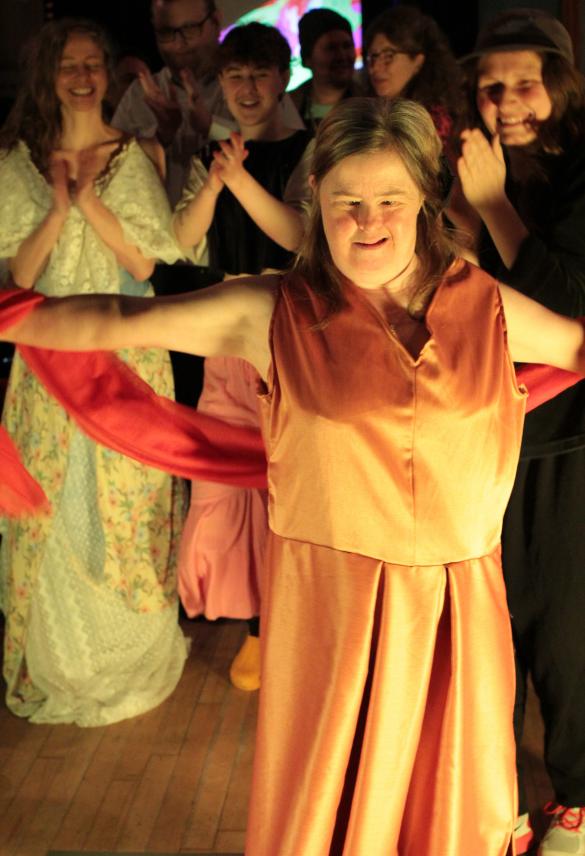 A person is standing in the front of the photo with her arms outstretched smiling and looking proud. She is wearing a salmon pink sleeveless satin dress with a thin red shawl round her forearms. In the background there are people in beautiful dresses cheering and clapping for her.