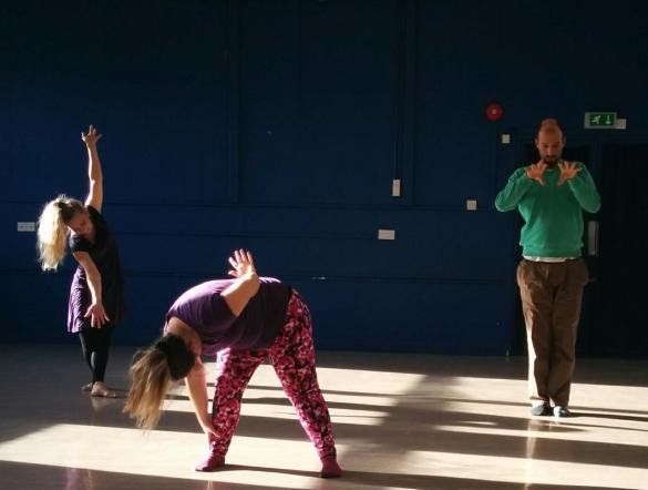 3 people dancing in a large hall. The person at the front and in the centre is standing, bent over with one hand holding their leg and the other resting on their back. The person the the back on the left is standing with one arm in the air and the over to the floor, they are looking at the floor. The person standing at the back right is standing with the hand held up to the chest and their hands stretched out