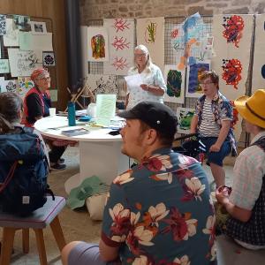 People seated at an exhibition with artwork on the walls around them. They are looking at a person who is standing up reading a poem from a piece of paper.