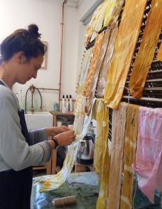 Naturally dyed strips of fabric are hanging up to dry. A person is holding the end of one of the lengths of fabric. The colours are lovely soft yellows, oranges and browns.