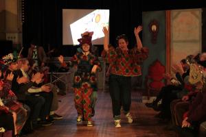 Two people in a fashion show wearing their costumes and waving their hands. The audiences is seated to the left and right of them and the background is dark.