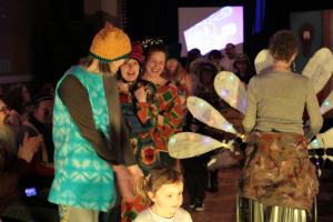 Artists in a fashion show on the catwalk smiling and sharing their costumes. The person on the left is wearing a turqoise dress and an orange hat.