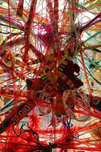 The image show brightly coloured wool, laces and threads woven and tied together on a frame, making a mesh of colour and texture, lit by strong sunlight from behind.