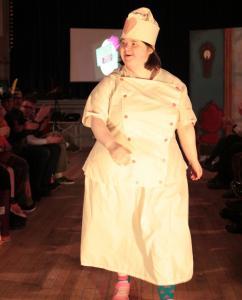 A person is walking down the catwalk at a fashion show wearing a white dress and hat with pink buttons and a pink heart on the hat. It is dark around her and there is an audience to her left and right.