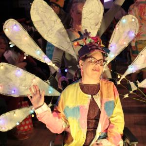 A person performing in a fashion show. She is seated and waiving her hand and smiling. Behind her there are big petals made of tissue paper lit up with fairy lights.