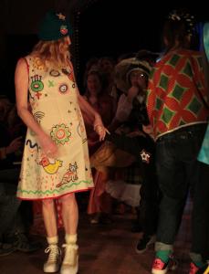 A person is on the left in the foreground wearing a cream coloured shift dress painted with beautiful bold patterns, a sun, a cat and a bird. She is wearing a black knitted hat. She is turning away to look at someone behind her in a darker space.