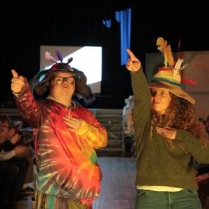 Two people are on the fashion show catwalk. They are both pointing with their right hands and wearing hats with feathers in.