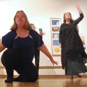 Three people art dancing in front of pictures on the wall in an art gallery. In the middle a person is crouching with their arms stretched out behind them. On either side is a dancer standing.