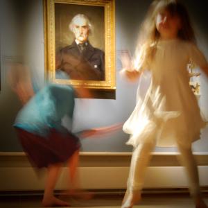 A young person in white slightly out of focus dances in front of a framed portrait in an exhibition. A blurred figure dances behind them.
