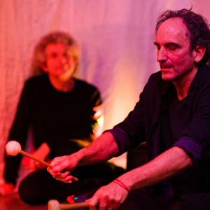 A person is sitting on the floor playing a drum with felt drumsticks. Another person is sitting down watching in the background