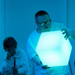 A person is holding a large light cube and another person is standing next to them watching them. A third person is sitting on the right of the image