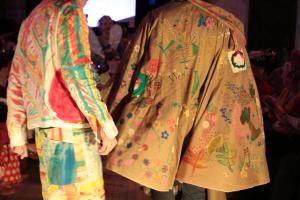 The photo shows the backs of two people but not their heads or feet. The person on the left is wearing a jacket and shorts hand-painted in pastel colours and the person on the right is wearing a light brown cape decorated with painted patterns, flowers and leaves. 