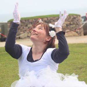 A dancer is dancing seated on the grass. They are wearing a white tutu dress and white gloves. Their hands are raised and they are smiling.