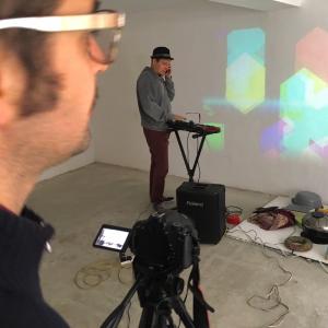 Two people are in a white space. One in the foreground is filming with a camera on a tripod and one in the background is making music on a keyboard. There is coloured light on the white wall.