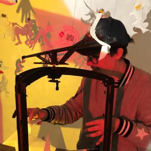 A person is moving shadow puppet figures around and in the background shadow puppet figures make shadows on the wall in yellow and red coloured light