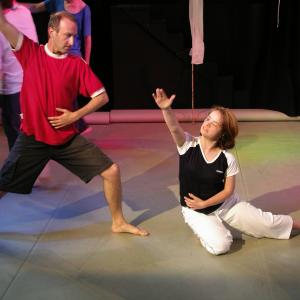 5 people dancing on stage. The 2 people in the front are most prominent. The figure at the front left is standing with wide legs and one arm raised in the air. The person at the front right is dancing on the floor with their left leg pointed towards the right and their right arm raised above their head.