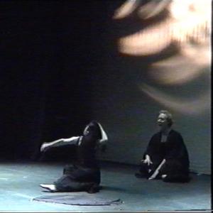 2 people performing on a dark stage. They are sitting on the floor and wearing black. 