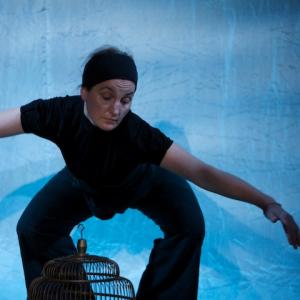 a person dancing on a blue stage. They are wearing all black. They are bent forward with their arms raised to the side