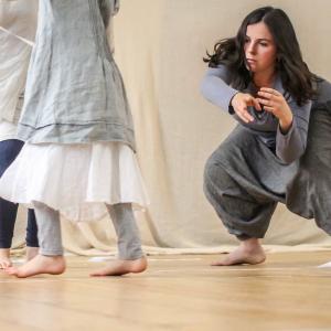 Three people are dancing in an indoor space. One is crouching with their arms forward gesturing. They all have bare feet and are wearing blue-ish grey linen clothing. The room is well lit with natural light.
