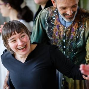 Two dancers in costume are smiling as others clap at the end of a performance. One is wearing black. The other is in a deep green robe with jewels and sequins on the front.