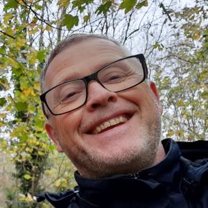 A photo of Nick's head and shoulders smiling down at the camera. There are trees in the background and Nick is wearing glasses.