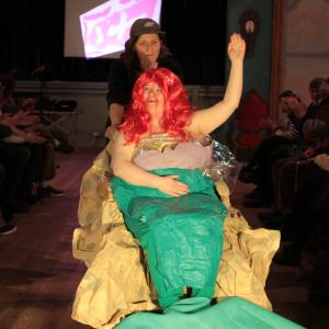 A person in a fashion showing wearing a mermaid costume including a pink wig. She is waiving with her left hand.