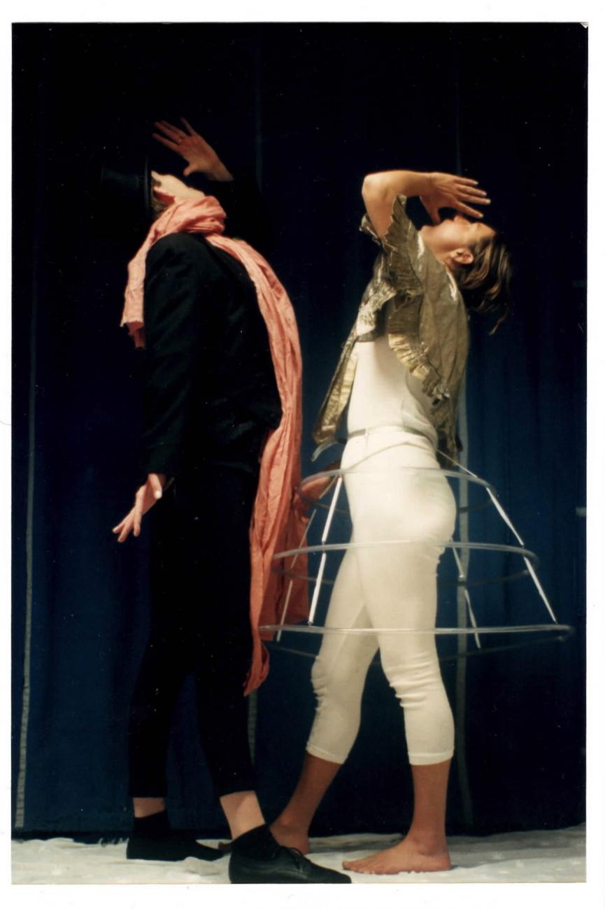 2 people performing. The both have their hands raised to their face and their heads are looking upwards. The person on the left is wearing black with an orange scarf. The person on the right is wearing a metal skirt structure over all white.