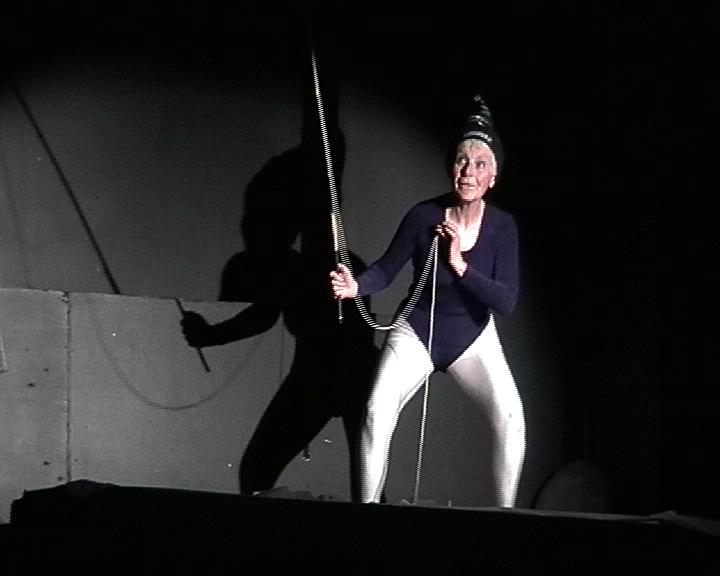 A person performing on stage. They are wearing a black leotard and silver leggings with a black hat. They are holding a sword with a chain attached