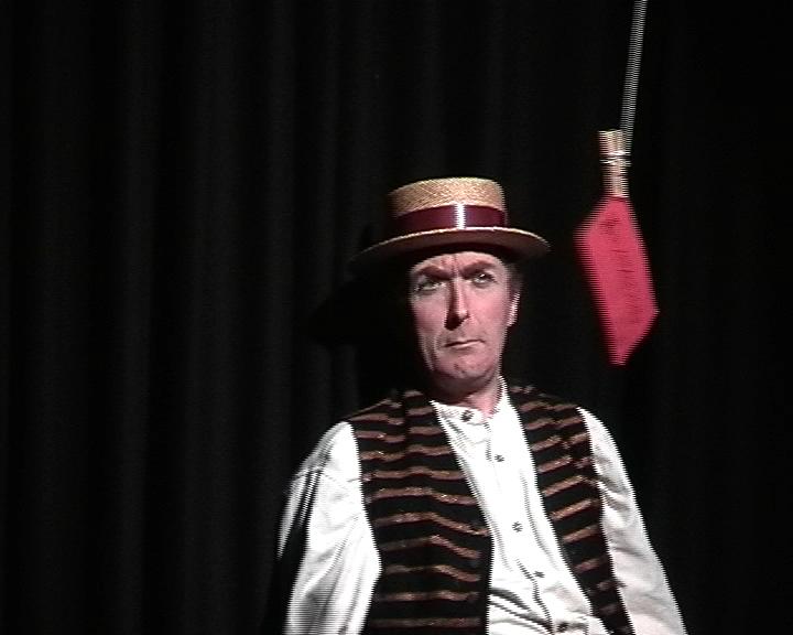 a person performing on a black stage. They are wearing a stripy waistcoat and a straw hat. There is a red envelope hanging from some string in front of them 