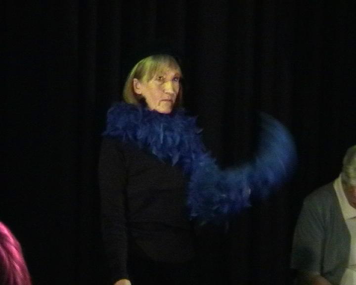 a person standing on a black stage. They are wearing all black with a blue feather bower. 