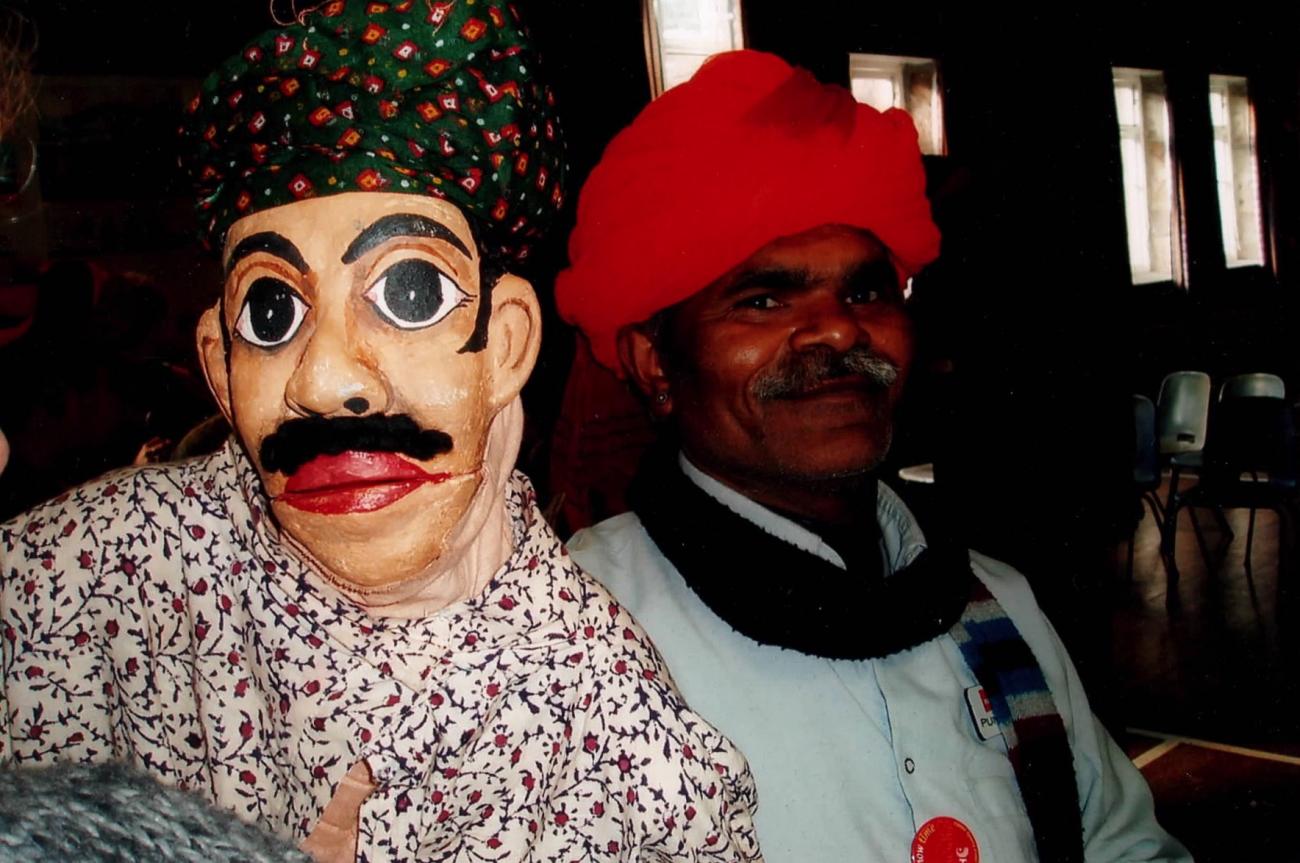 A figure holding a puppet. The figure is wearing a red turban and smiling at the camera. The puppet is wearing a green and red spotted turban and has big eyes, a black moustache and red lips. 