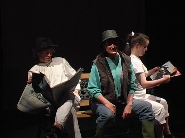 3 people performing on stage in costume. They are sitting on a bench. 2 people are reading a newspaper and one is sitting looking out towards the audience.