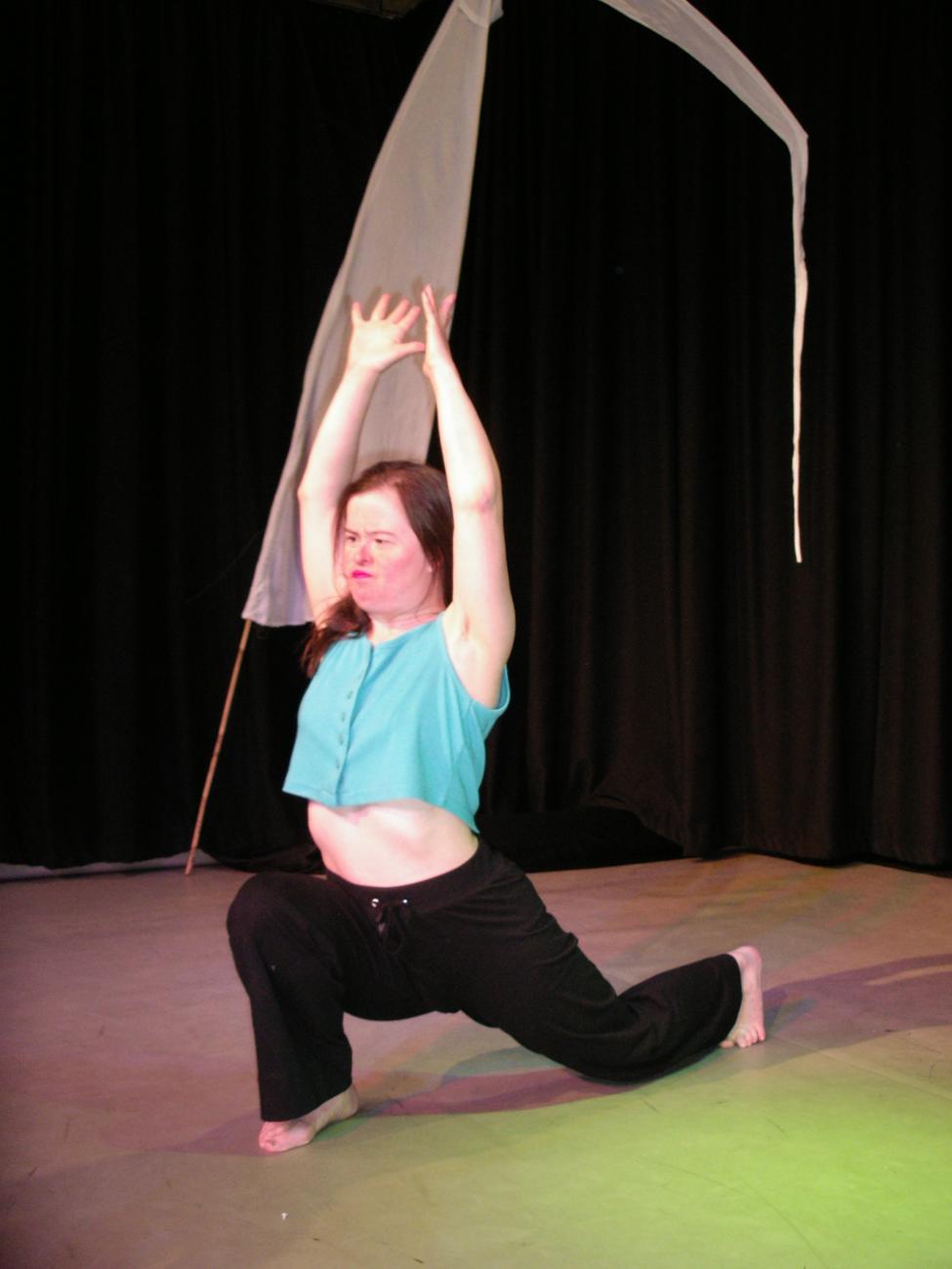A figure in black trousers and a blue top dancing on stage. They are standing in a lunge position. They have both their arms raised above their head. 
