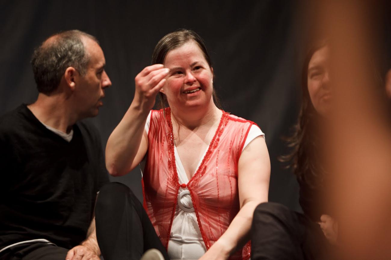 3 people sitting together. The person in the centre is wearing a white dress with a red waistcoat. They are smiling and reaching their hand towards their face. The person on the right is slightly covered by a blurry object but you can see that they are wearing black and looking at the camera. The person on the left is wearing black and looking ay the person in the middle. 