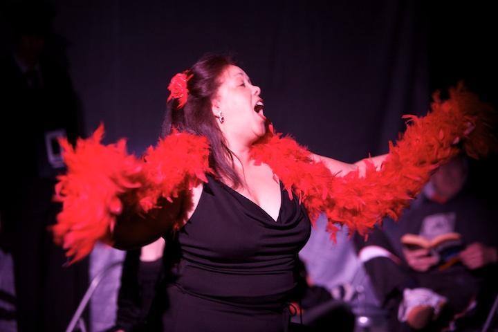 A person in a black dress and red feather bower singing on stage.