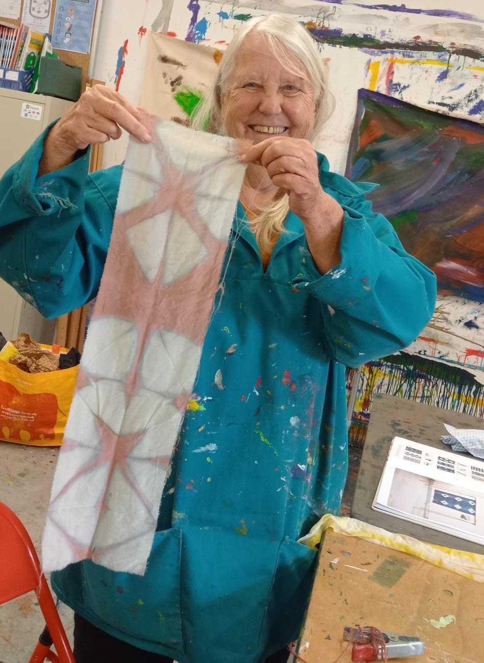 A person is holding up a length of fabric which they have dyed themselves. The fabric is a soft pink with white diamond shapes and brown lines. The person looks happy and proud